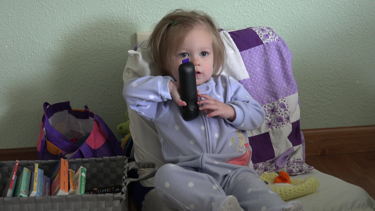 The child remains seated in the Westminster, Colorado interior, looking forward while gripping the black remote control in front of a pale green wall and quilted chair back.