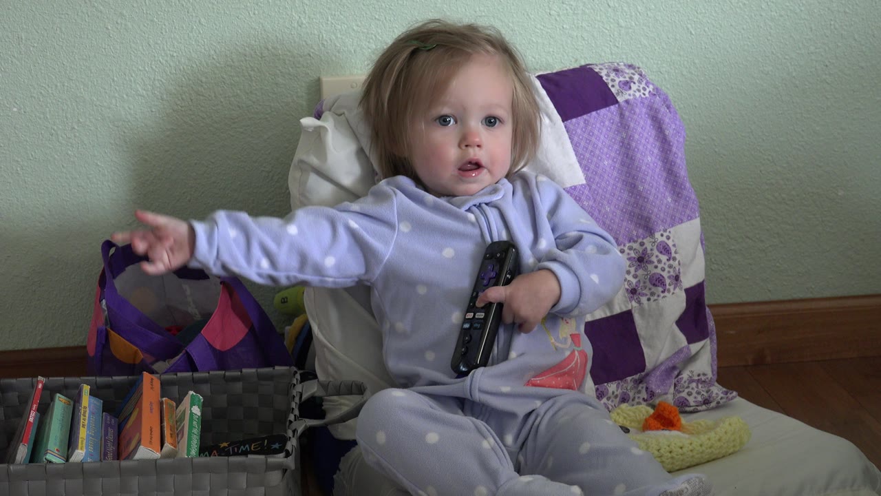 Young girl sits in a chair holding a remote and reaching toward a media basket.