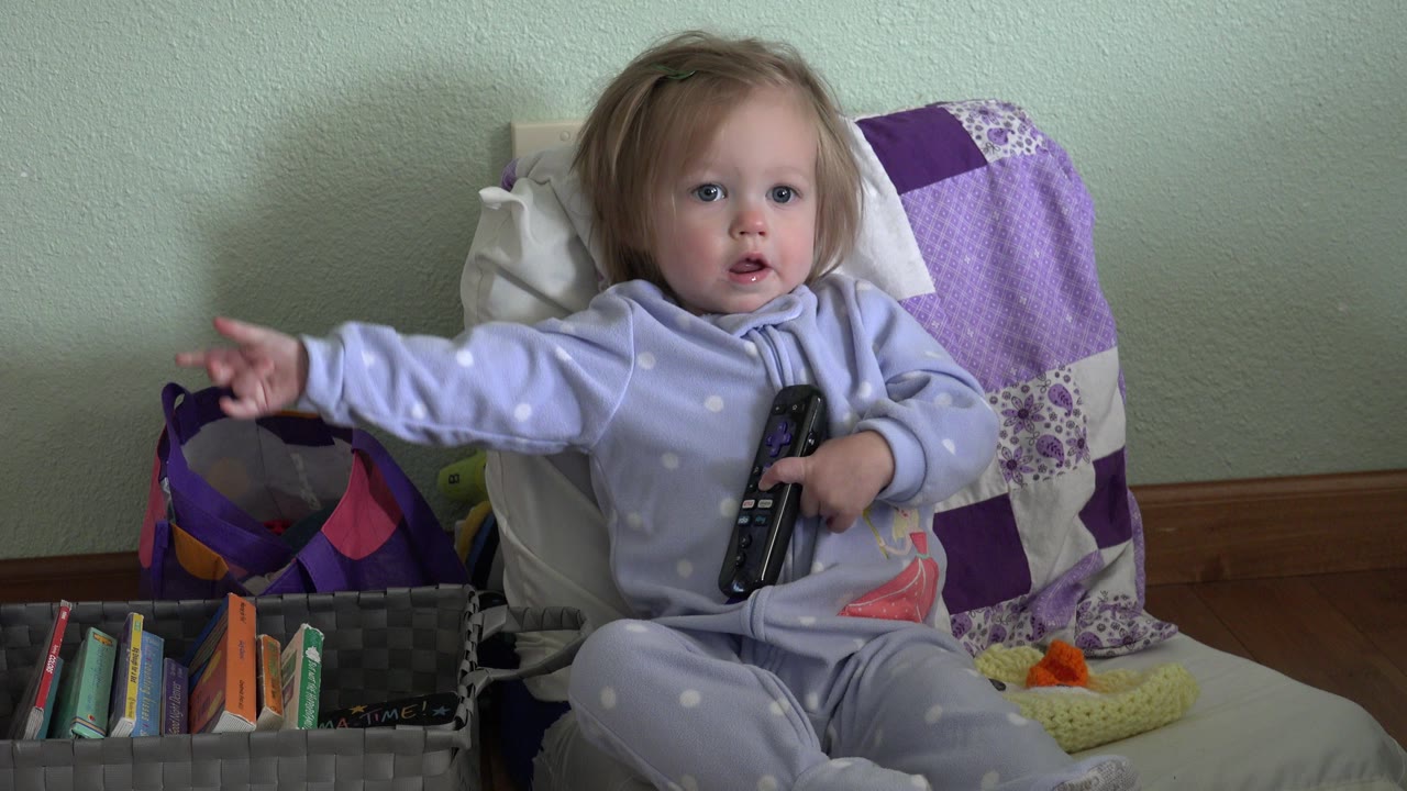 In Westminster, Colorado, the child looks forward while holding the remote against her body and pointing her outstretched hand toward the media basket beside the chair.