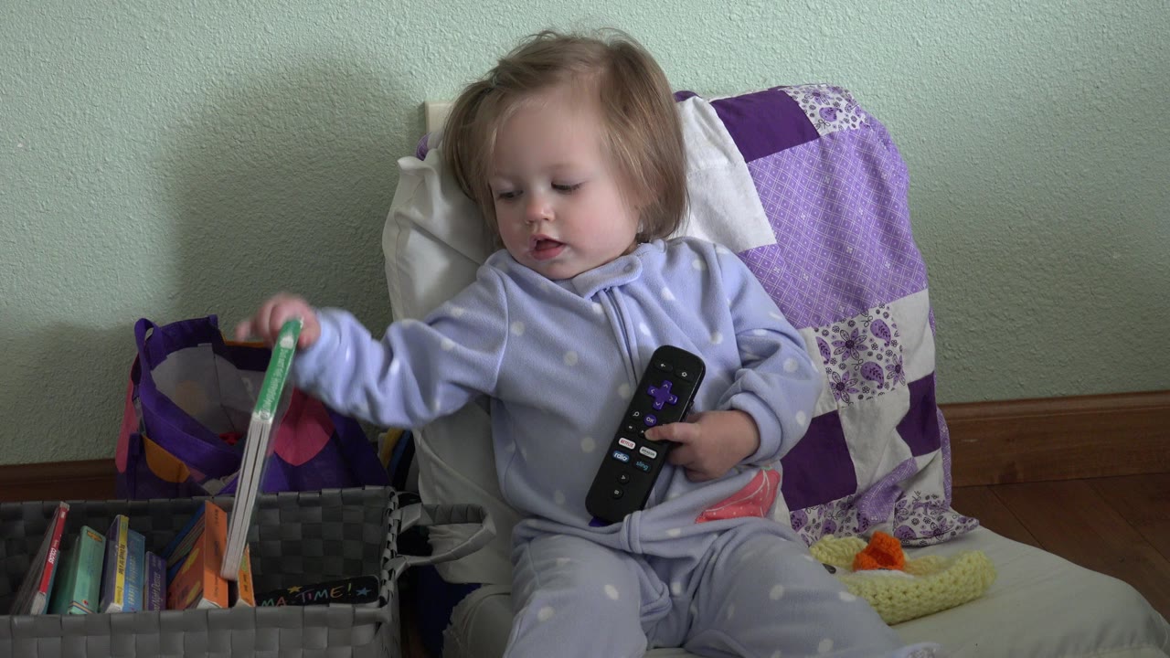 Toddler sitting with a remote while reaching toward books.