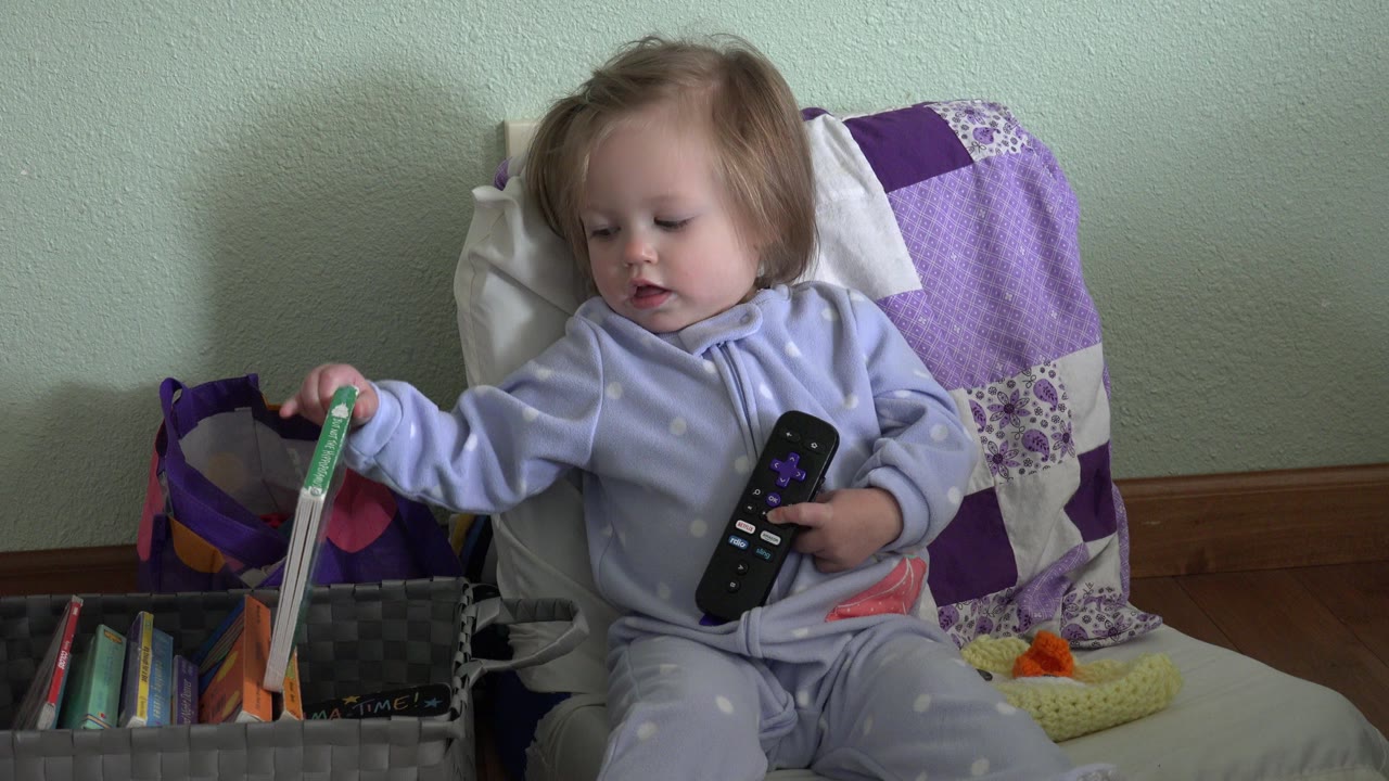 In Westminster, Colorado, the toddler remains seated, holds the remote against her pajamas, and touches a green children's book in the bin at her side.