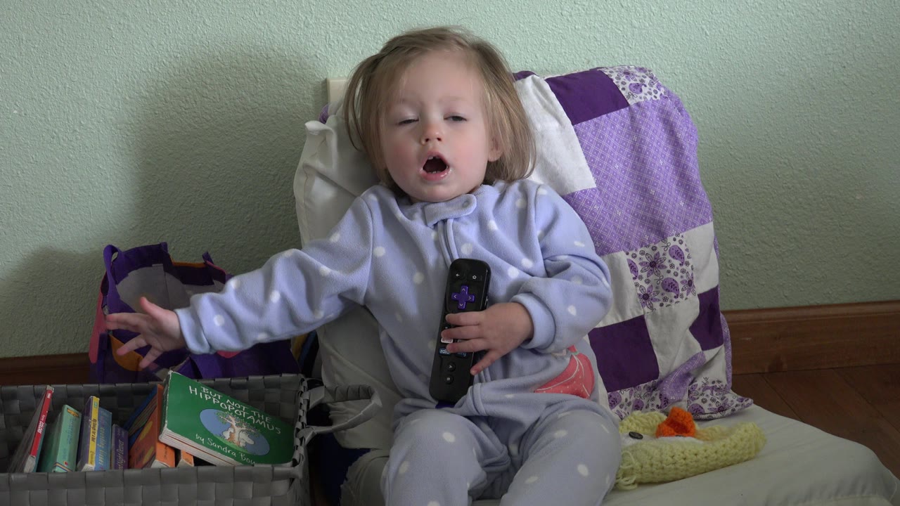 A young child in Westminster, Colorado sits in a cushioned chair, holds a remote control, and reaches toward a basket of books at the start of the clip.
