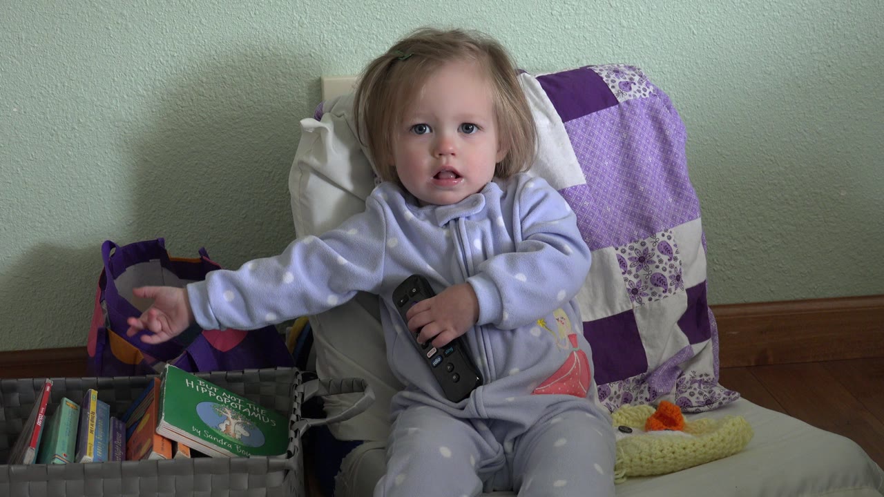 Toddler sitting on a chair holding a remote beside a bin of books.
