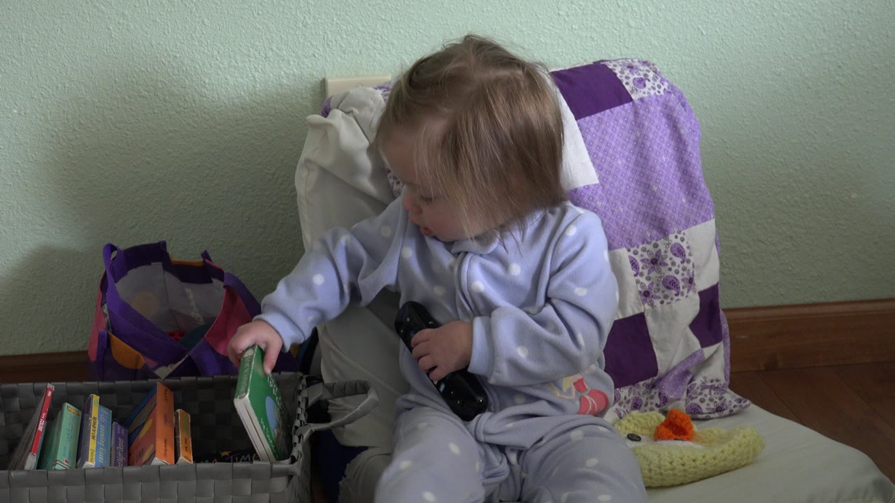The toddler looks down at a green book near the front of the gray bin, with colorful book spines and a purple blanket behind the chair.