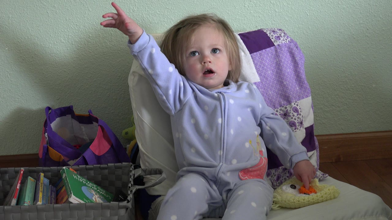 Young child in pale pajamas reaches upward while sitting on a couch.