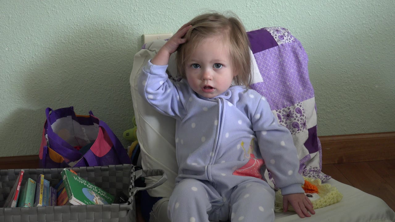 Toddler girl seated in a child chair with one hand on her head.
