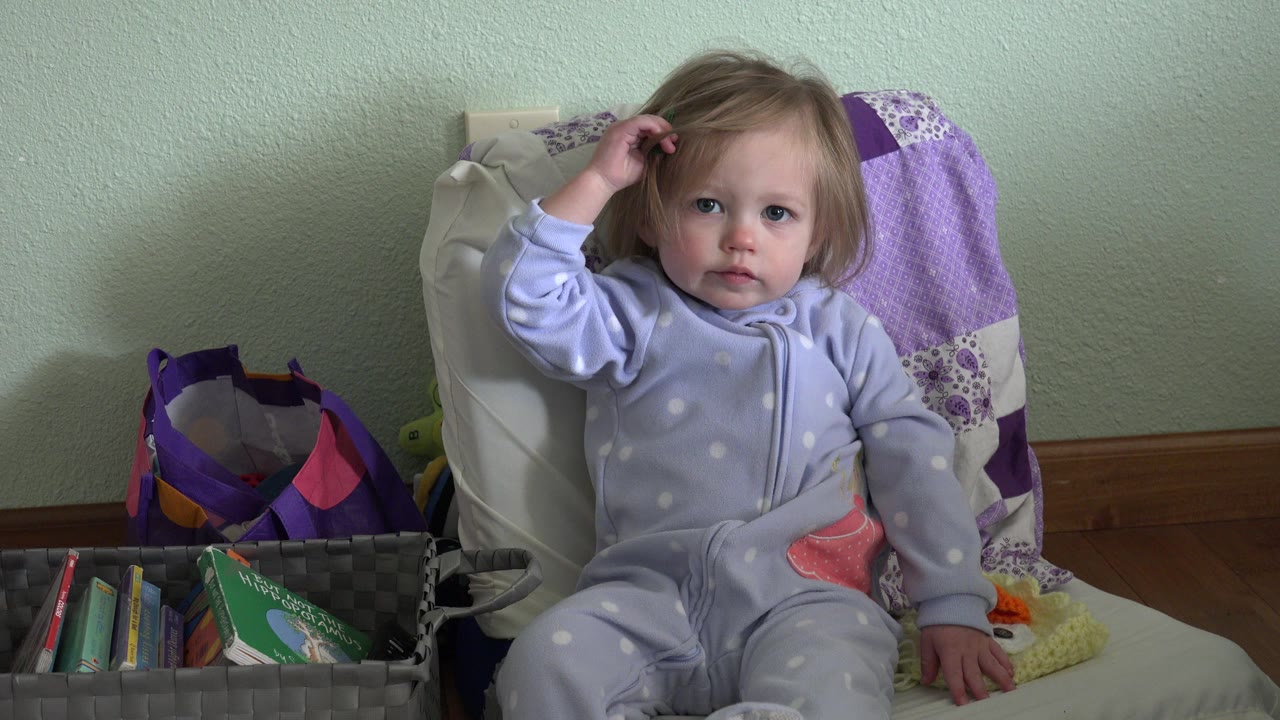 Toddler girl in pajamas touches her hair while seated indoors.