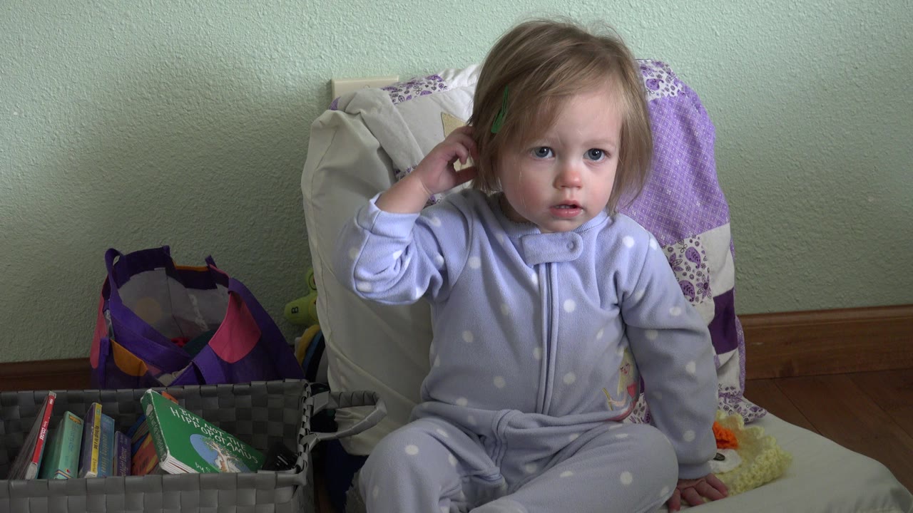 Young girl sitting on a bed with one hand near her ear.