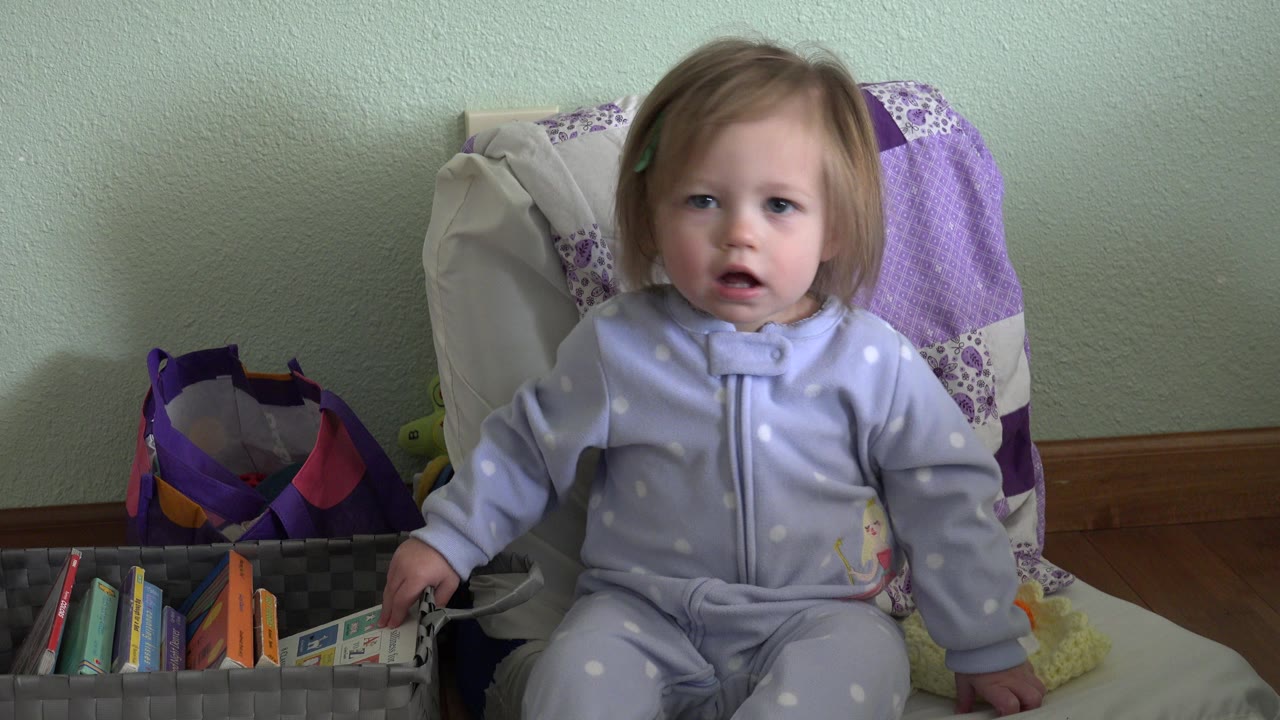 The toddler remains centered in the chair in Westminster, Colorado, facing the camera while resting a hand on the gray book basket.