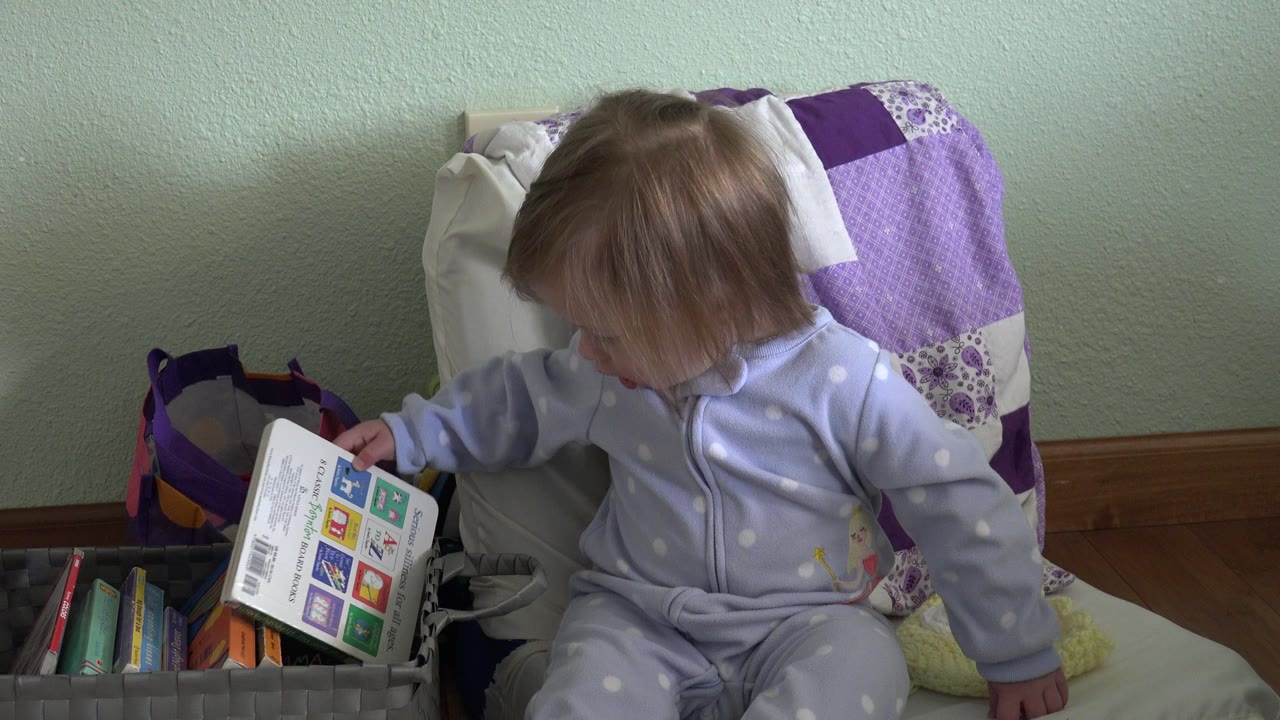 In Westminster, Colorado, the baby holds the book upright near the edge of the gray book bin while looking down toward it.