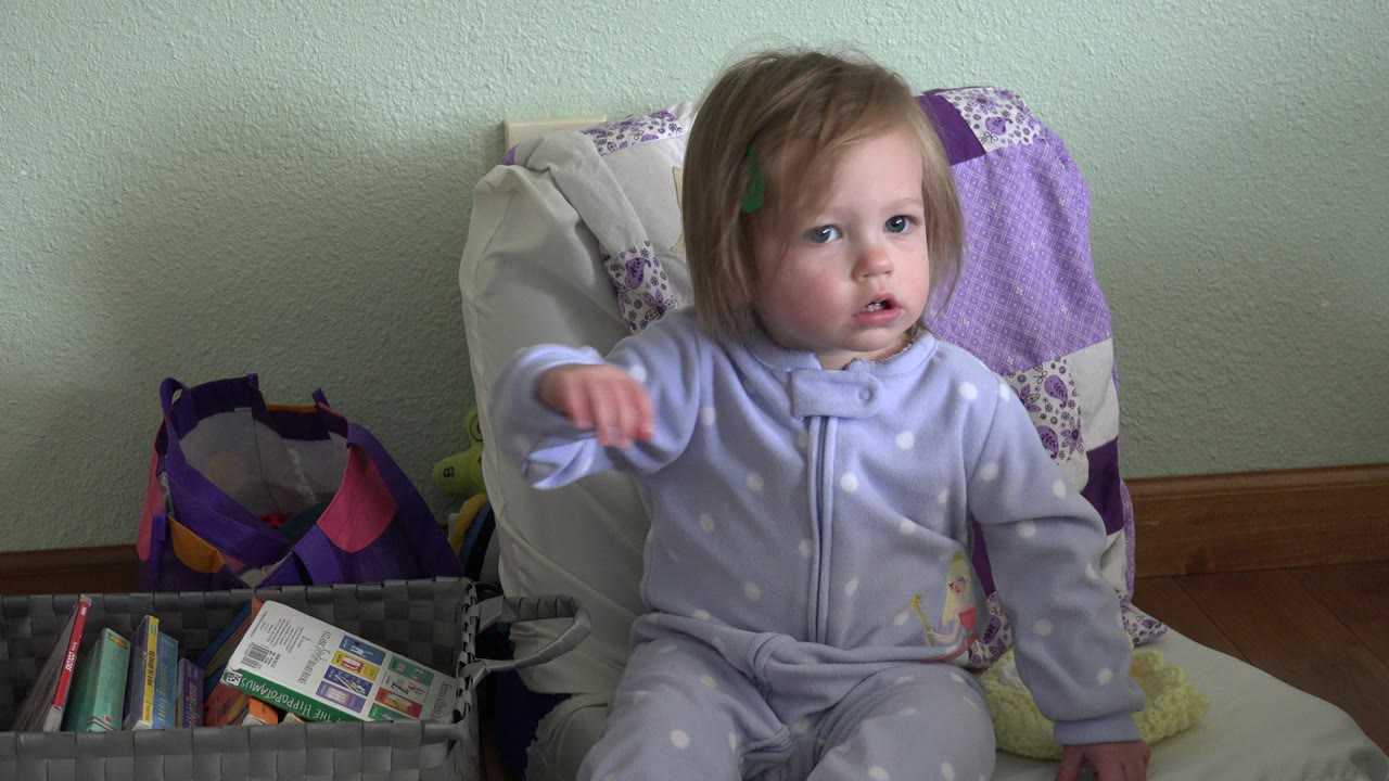 Toddler in purple sleeper seated on a chair beside a basket of books.