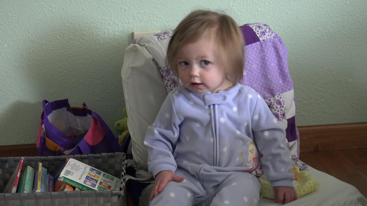 A toddler in Westminster, Colorado sits upright with hands near the lap, glancing slightly off camera while the purple chair cover and book basket remain in view.