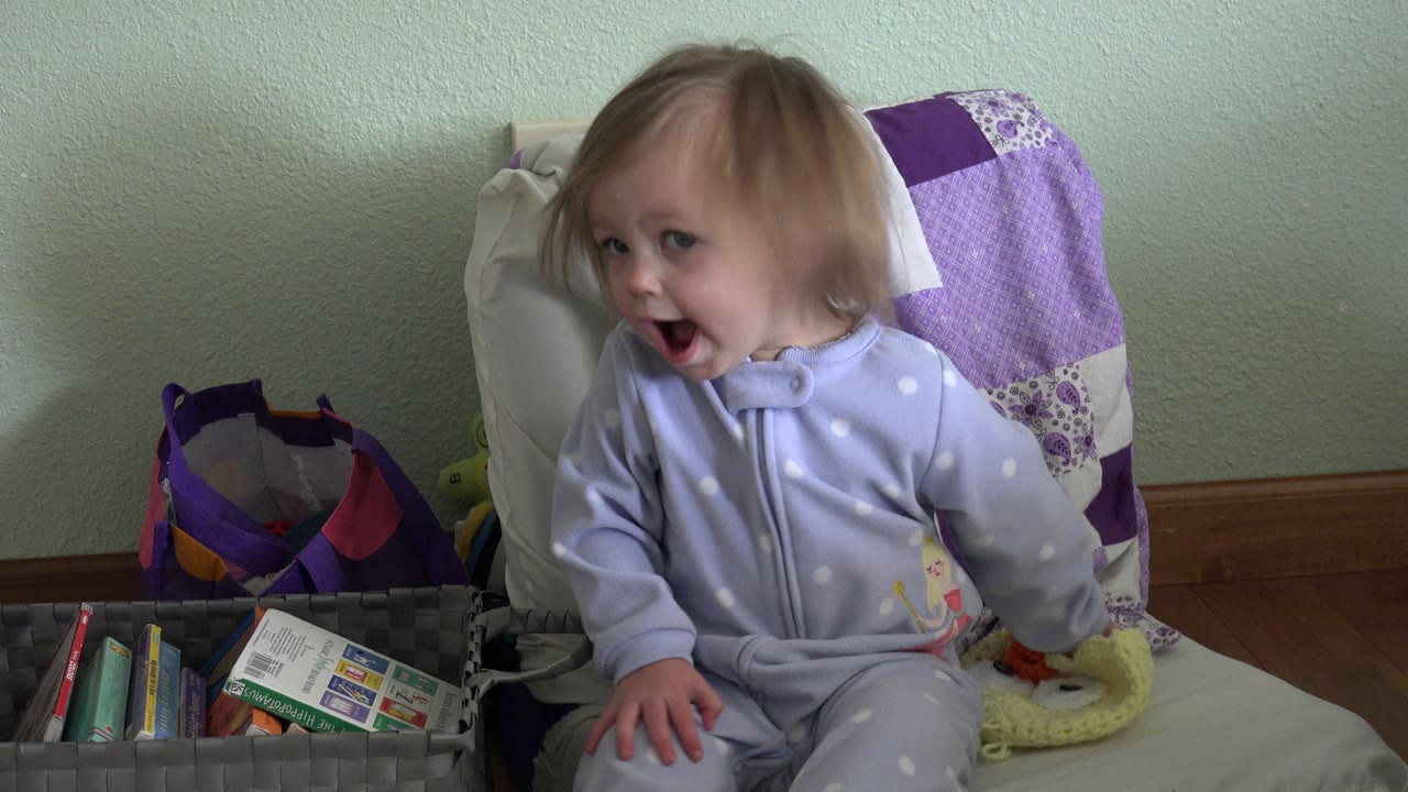 A baby in a pale sleeper sits on a cushioned chair in a Westminster, Colorado home setting, looking left with an open mouth beside a basket of toys and books.