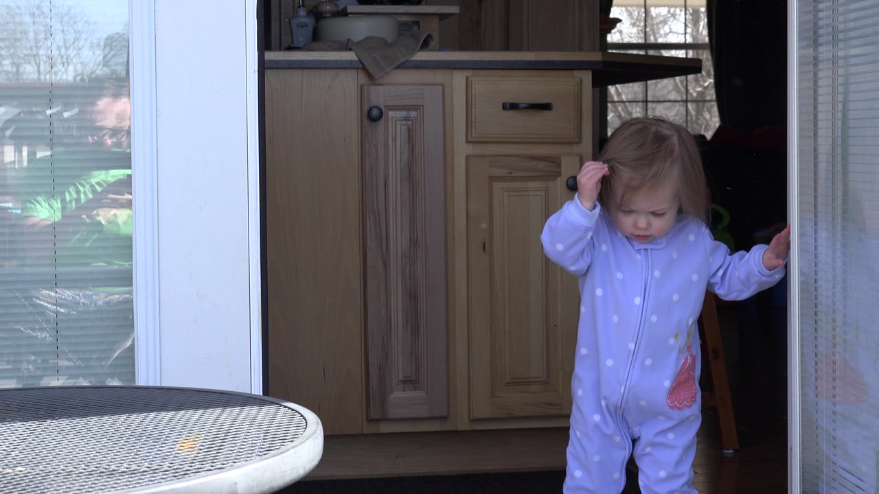 At 0:00 in Westminster, Colorado, a toddler in a light purple sleeper stands near an open glass patio door with one hand near her hair and a metal outdoor table in the foreground.
