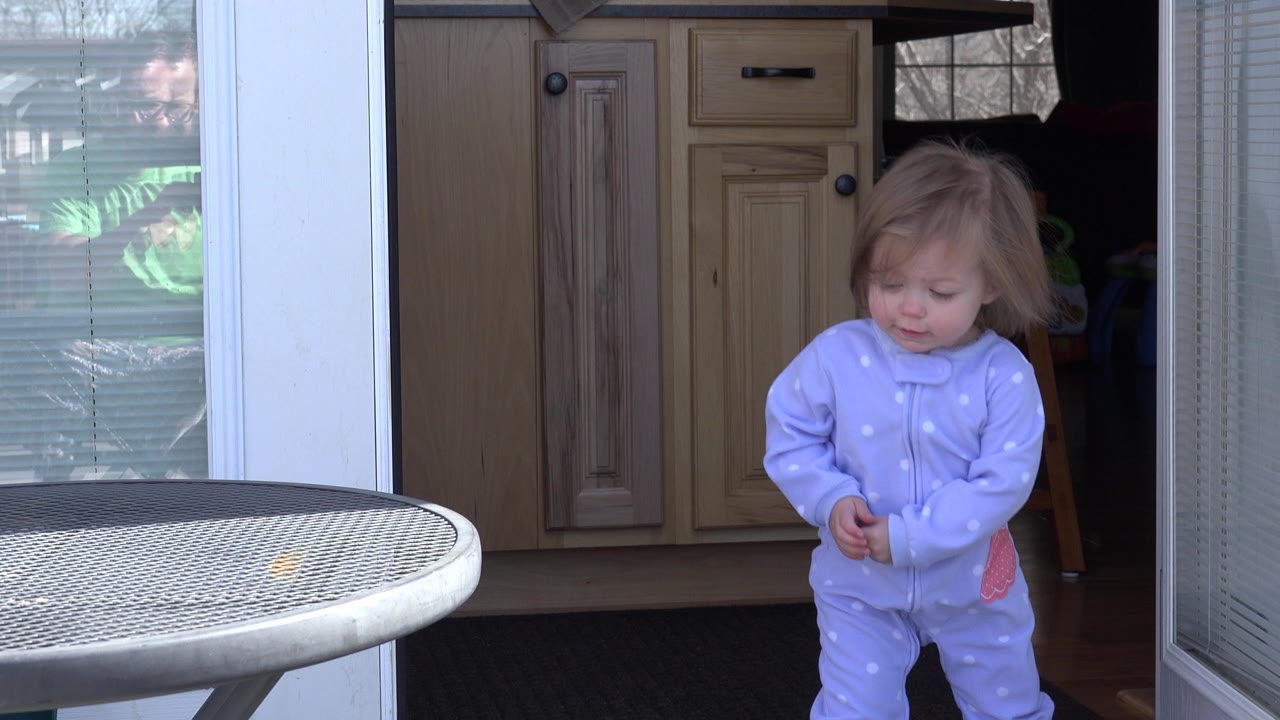 Toddler in a pale blue outfit standing by an open glass doorway.