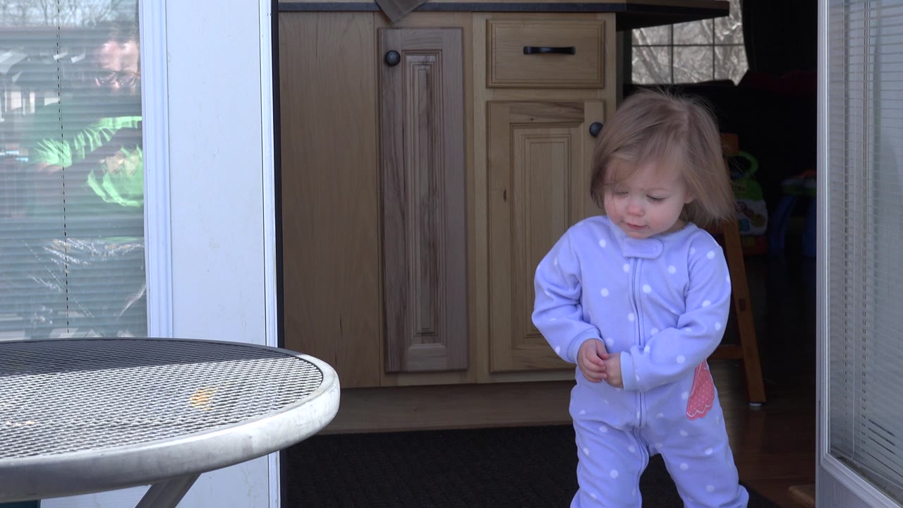 The toddler looks down while standing near the open doorway, with wood cabinets visible inside the Westminster home and the patio table edge in front.