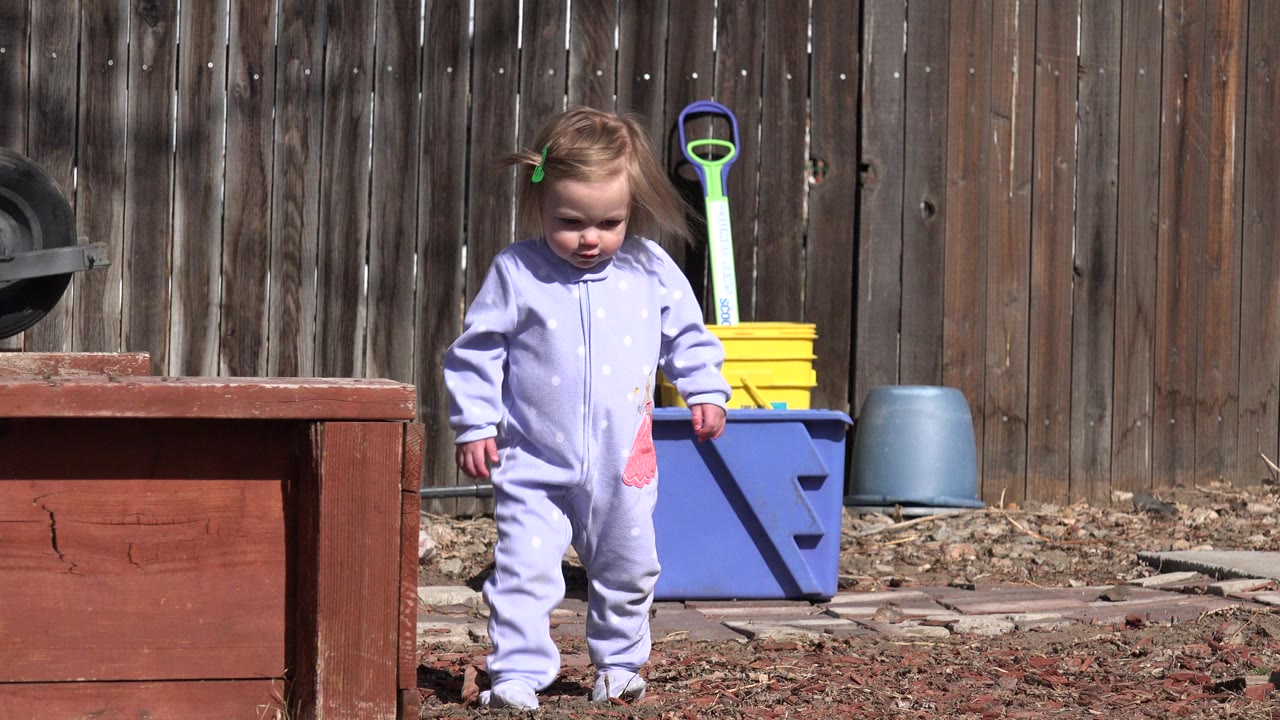 Toddler in a pale purple onesie standing in a backyard near a red wooden planter.
