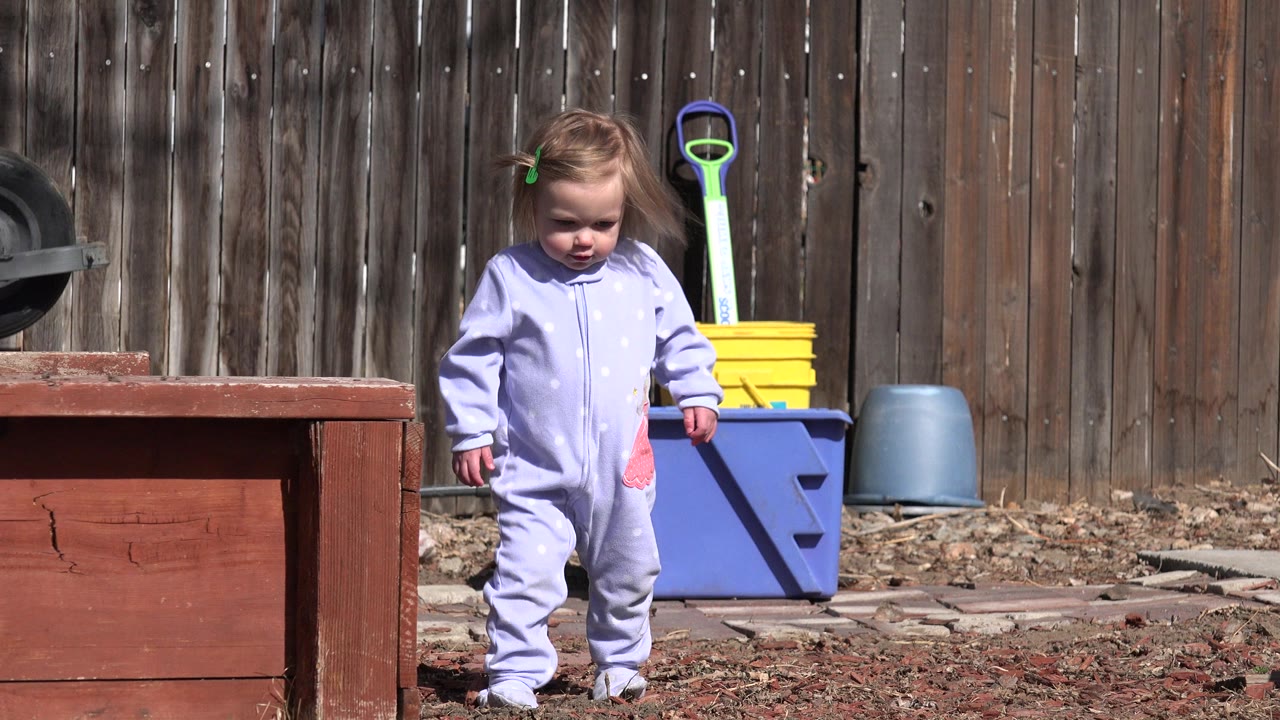 A toddler in a pale purple onesie walks forward in a Westminster, Colorado backyard, framed between a red wooden planter and yard supplies against a wood fence.