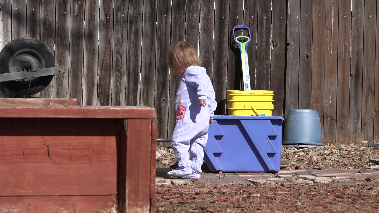 The child continues walking across the fenced Westminster backyard, passing the blue bin and yellow bucket while the red wooden structure blocks part of the lower frame.