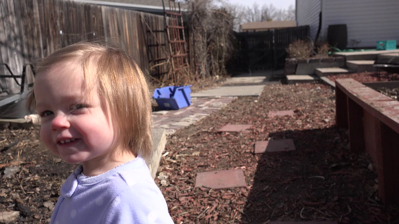 A blond toddler smiles near the camera in a sunny Westminster, Colorado backyard with a wood fence, mulch, stepping stones, and patio items behind them.