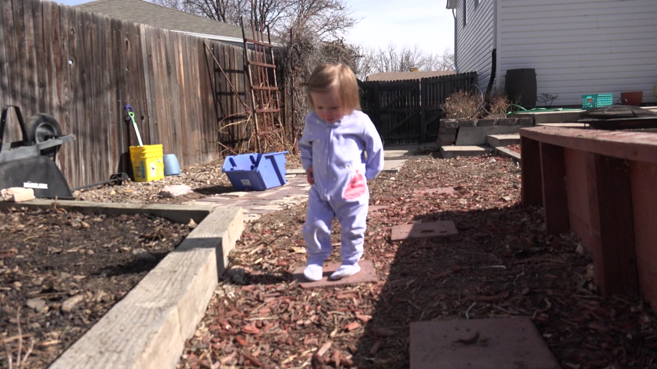 Toddler girl in purple standing on mulch beside a raised garden bed.