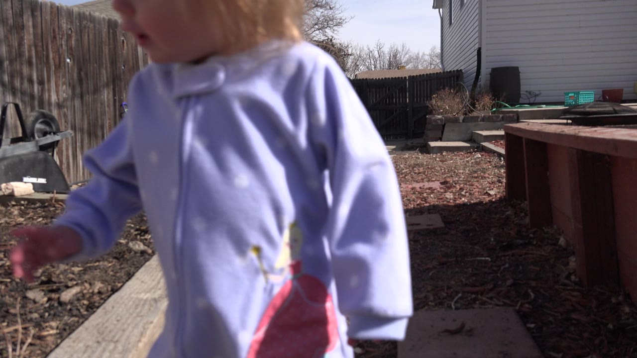The child remains in the foreground of the Westminster backyard, partially cropped at the face while the sunlit mulch bed and fence line stay visible behind her.