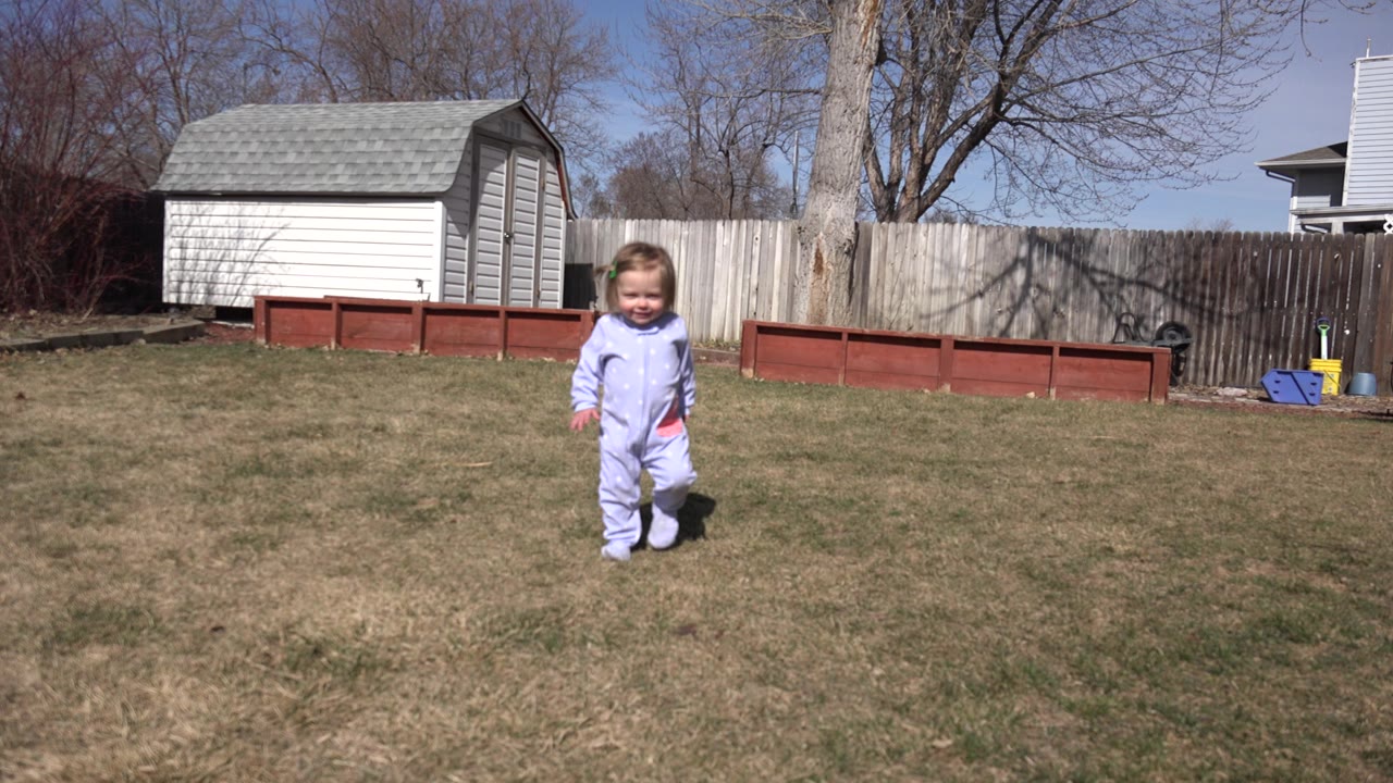 The child continues toward the camera in the Westminster backyard, still framed wide with the shed and fence stretching across the background.