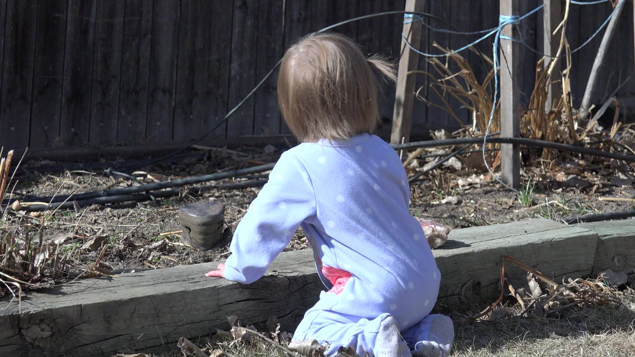 Toddler in blue pajamas sitting on backyard grass near a timber garden edge.