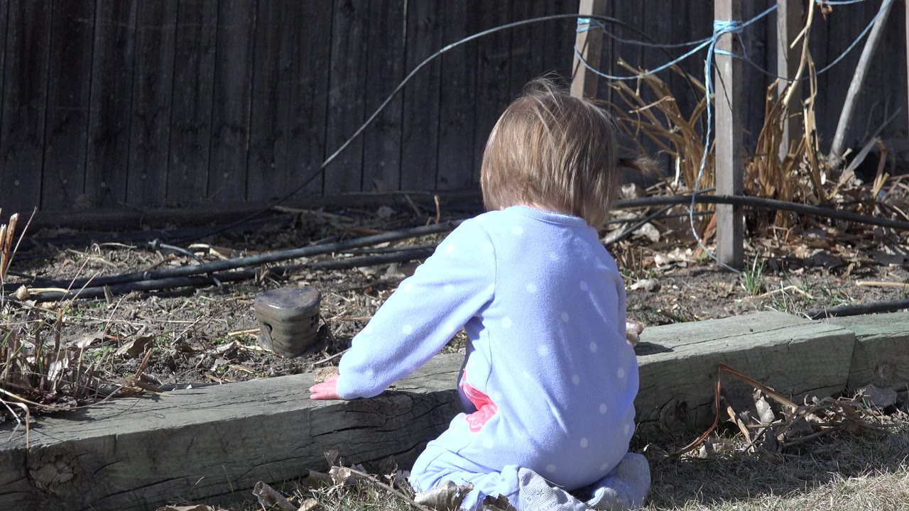 Toddler in blue outfit kneeling beside a wood curb outdoors.