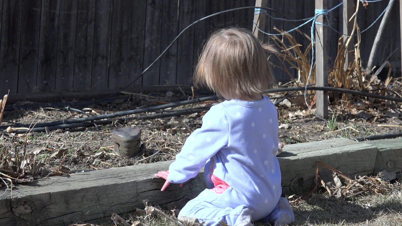 At 0:01 in Westminster, Colorado, the child remains turned away from camera near the timber edging, with dry yard plants and black tubing behind them.