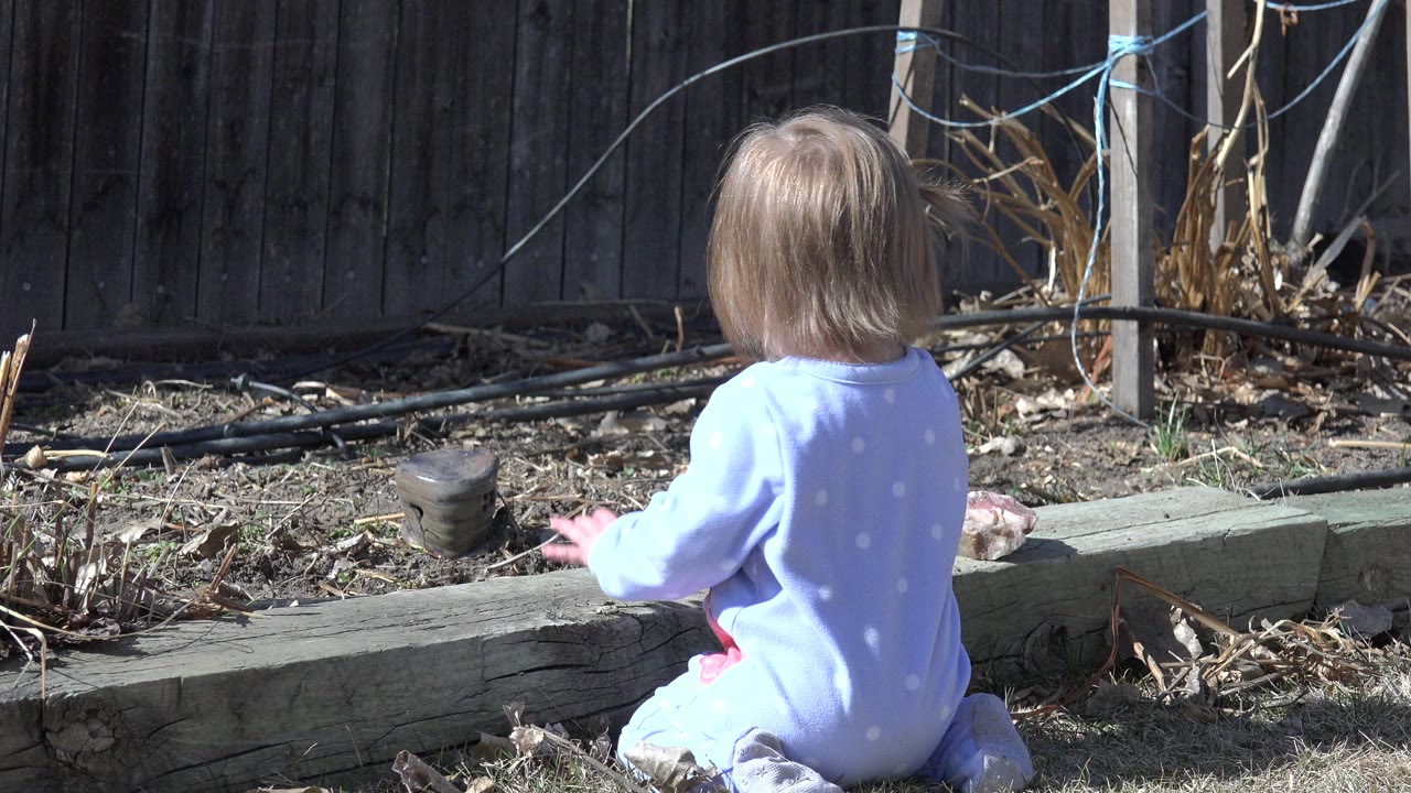 Toddler in blue pajamas sits beside a timber curb in a dry yard.