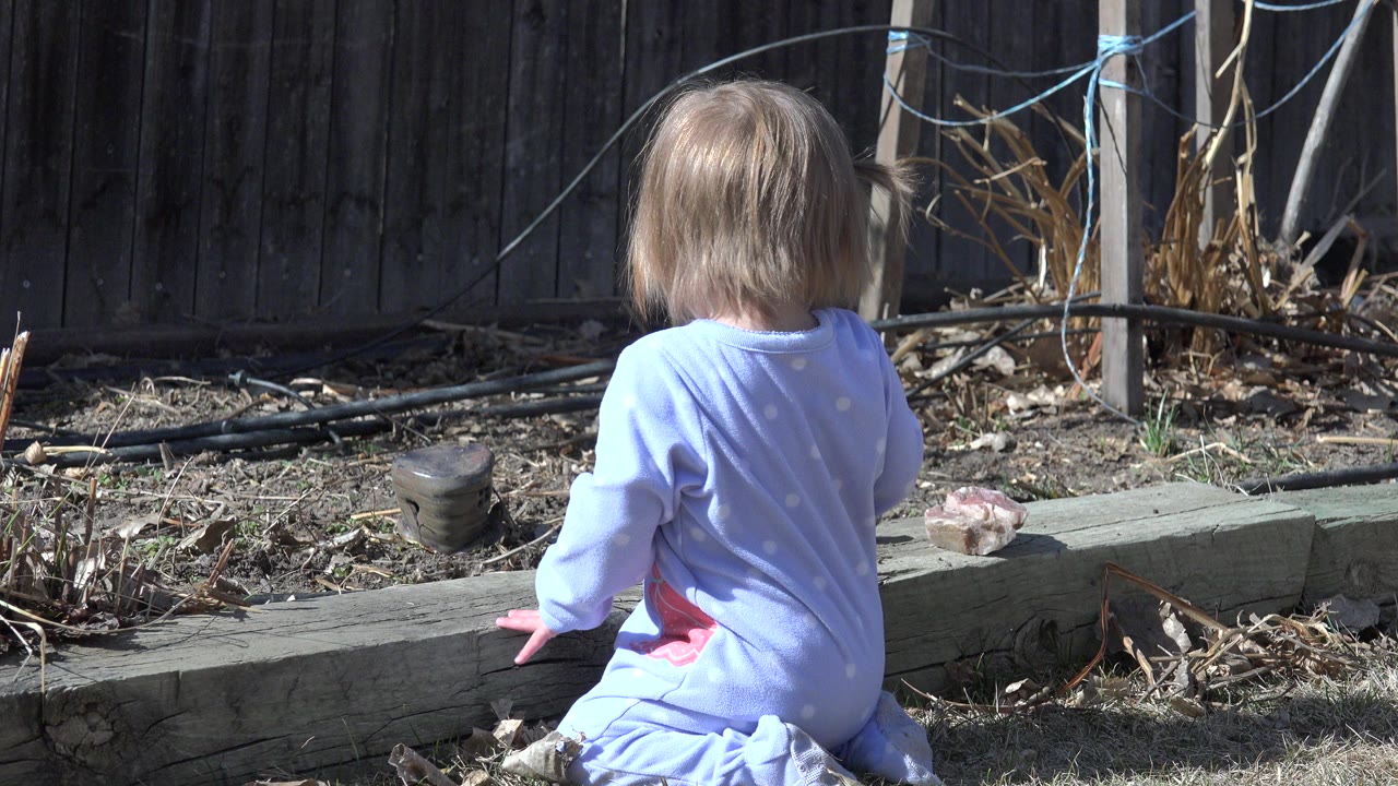 Toddler in a blue outfit sitting beside a garden bed