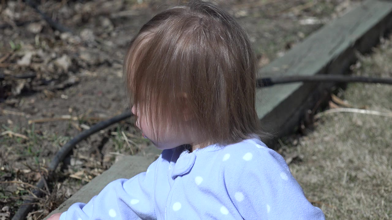 A young child with long brown hair in a light blue polka-dot top sits outdoors in Westminster, Colorado, turned left with dirt, dry grass, and a dark hose behind them.