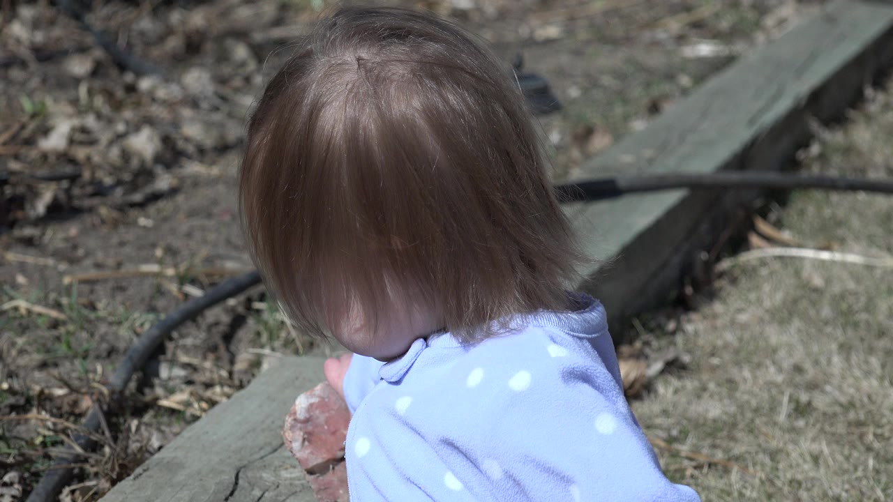 The child remains turned mostly away in Westminster, Colorado, with hair covering the face and a stone held near weathered wood and patchy grass.
