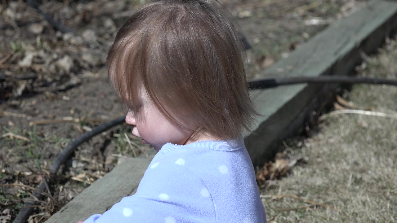 Blonde child in a light blue dotted shirt seen in profile outdoors in Westminster, Colorado, with dry grass and a green garden border behind them.