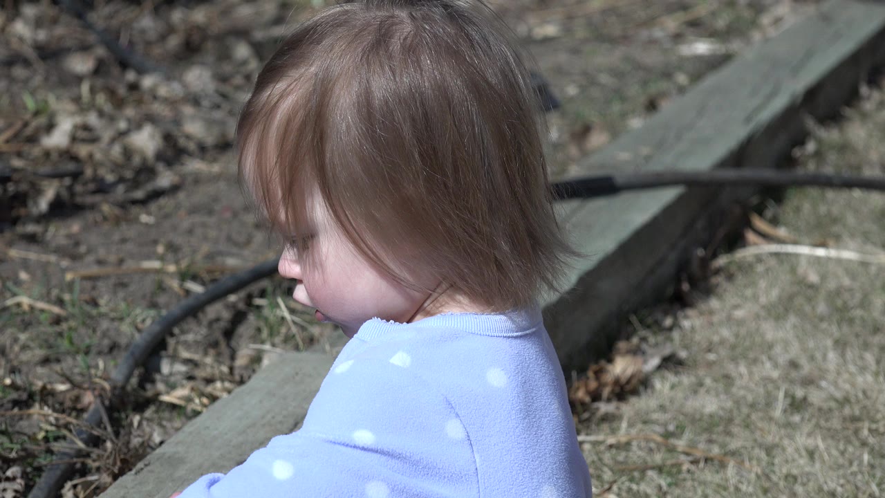 Young blonde child remains turned to the side in Westminster, Colorado, with hair covering part of the face and a black hose crossing the yard behind them.