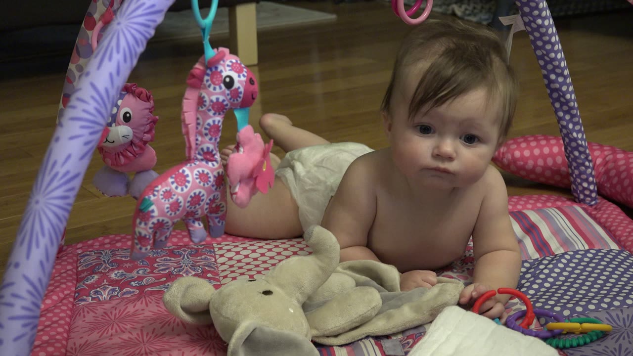 At 0:01 in Westminster, Colorado, the baby faces the camera more directly while lying between the hanging toys and colorful plastic rings.