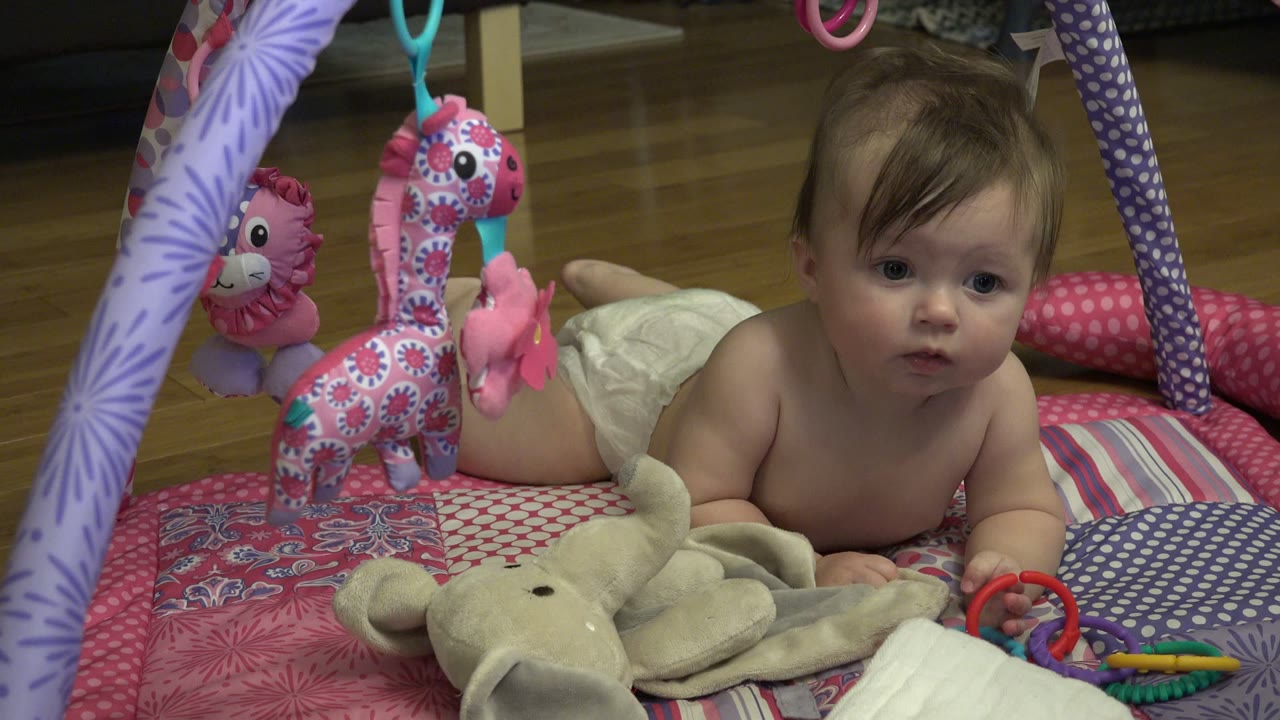 At 0:00, a baby girl lies on her stomach on a pink and violet baby gym in Westminster, Colorado, with a plush elephant and hanging toys around her.