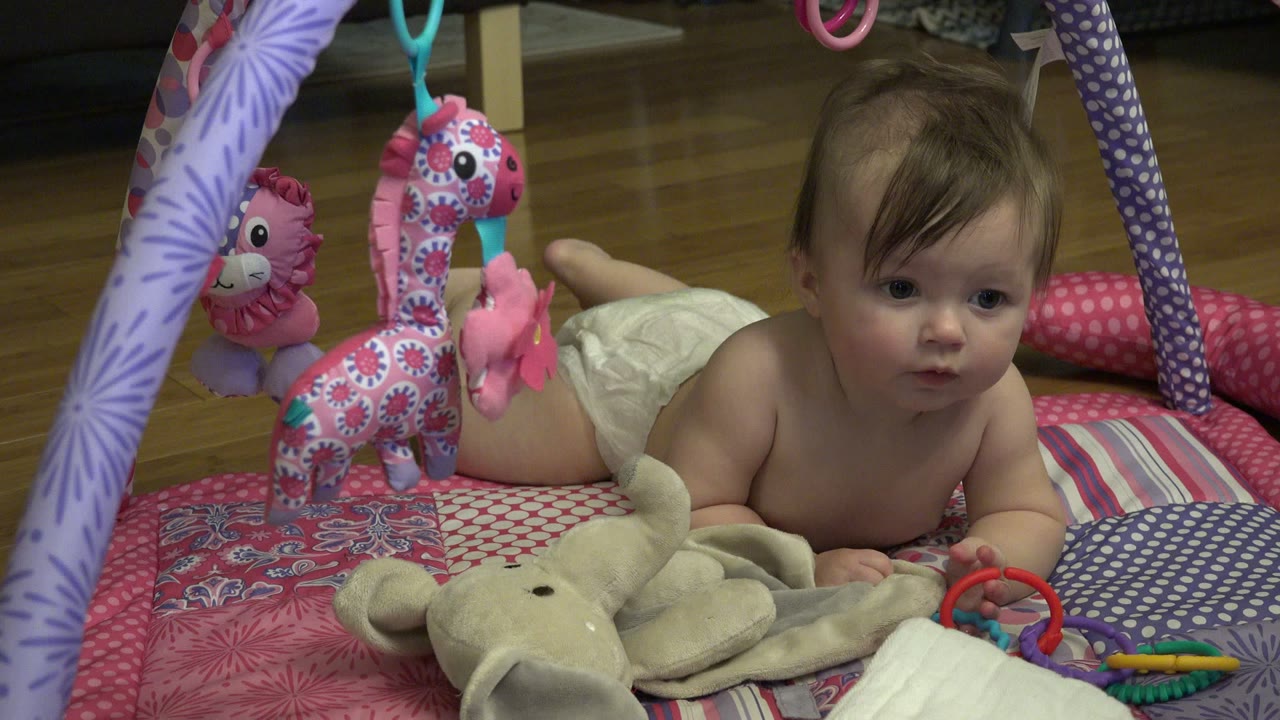 At 0:01, the baby girl remains on her stomach in Westminster, Colorado, looking forward beside colorful plastic keys and a beige plush elephant.