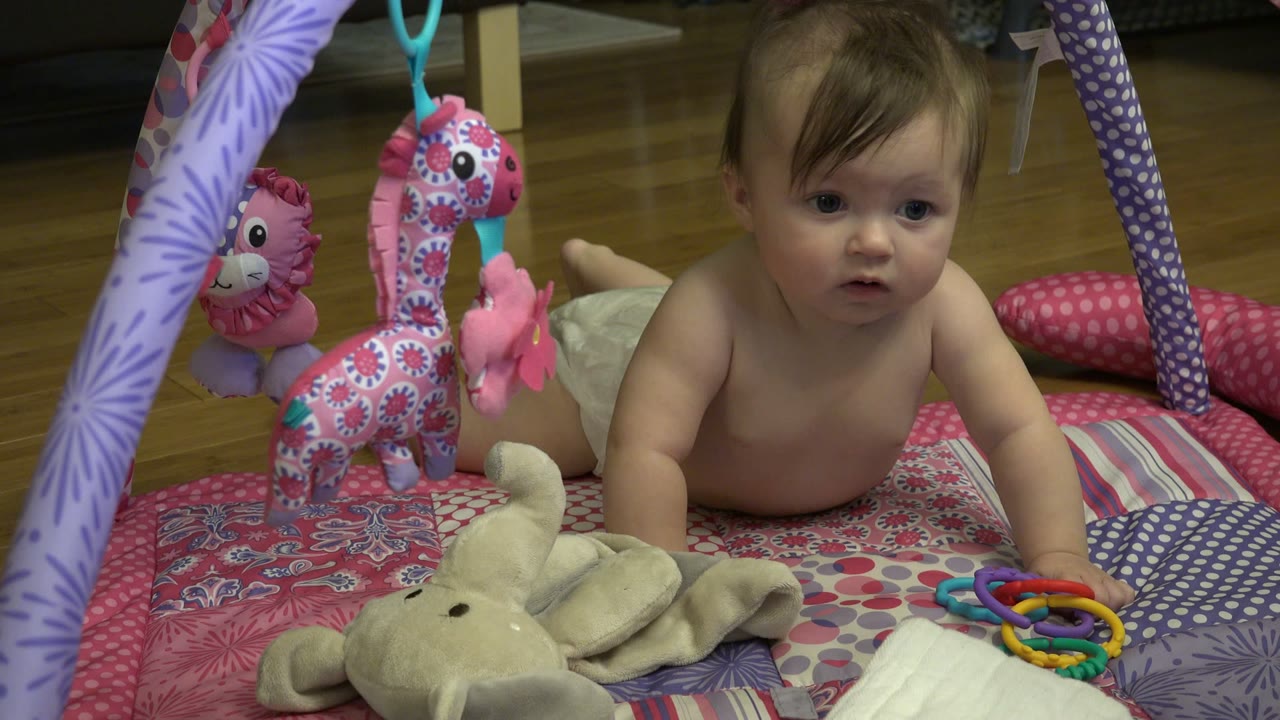 At the start of the Westminster, Colorado clip, a brown-haired baby lies on a pink and purple play mat with hanging plush toys and plastic rings nearby.