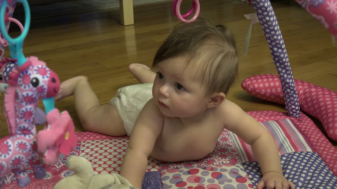 At the start of the Westminster, Colorado clip, a diapered baby lies on their stomach on a patterned play mat while pink and teal hanging toys fill the foreground.
