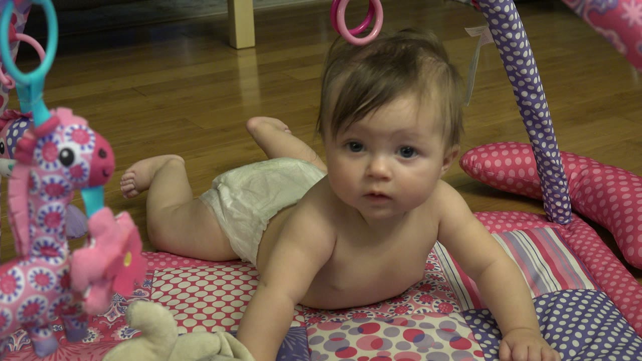 One second in, the baby remains on the Westminster home play mat, looking forward while colorful hanging toys partially frame the left side of the shot.