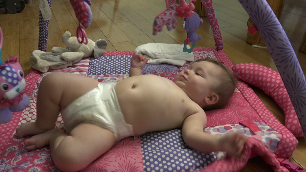 A baby in a diaper lies on a pink patterned play mat under dangling toys in a Westminster, Colorado home interior.