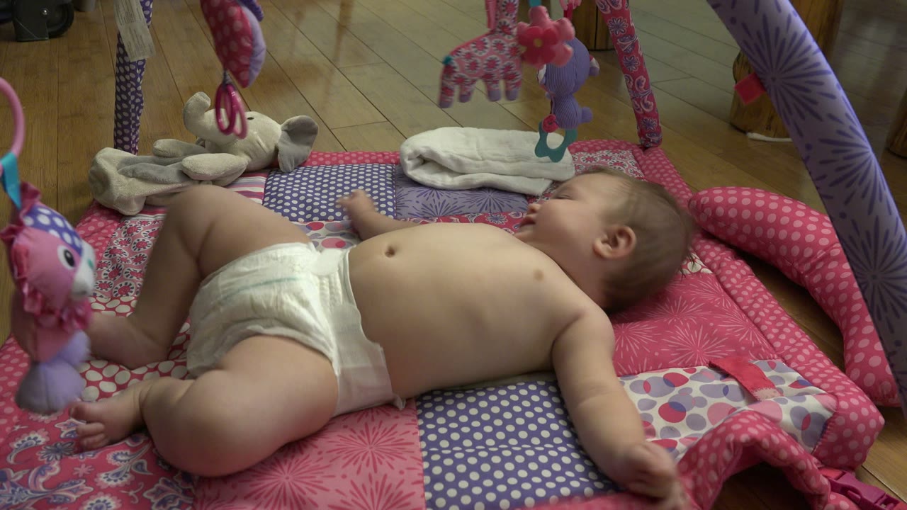 The baby turns slightly on the patterned mat, with one knee lifted and plush toys hanging above in the Westminster, Colorado interior.