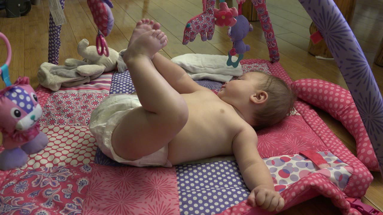 The baby remains on its back on the patterned mat, holding feet high while a stuffed toy and fabric arch hang above the play area in Westminster, Colorado.