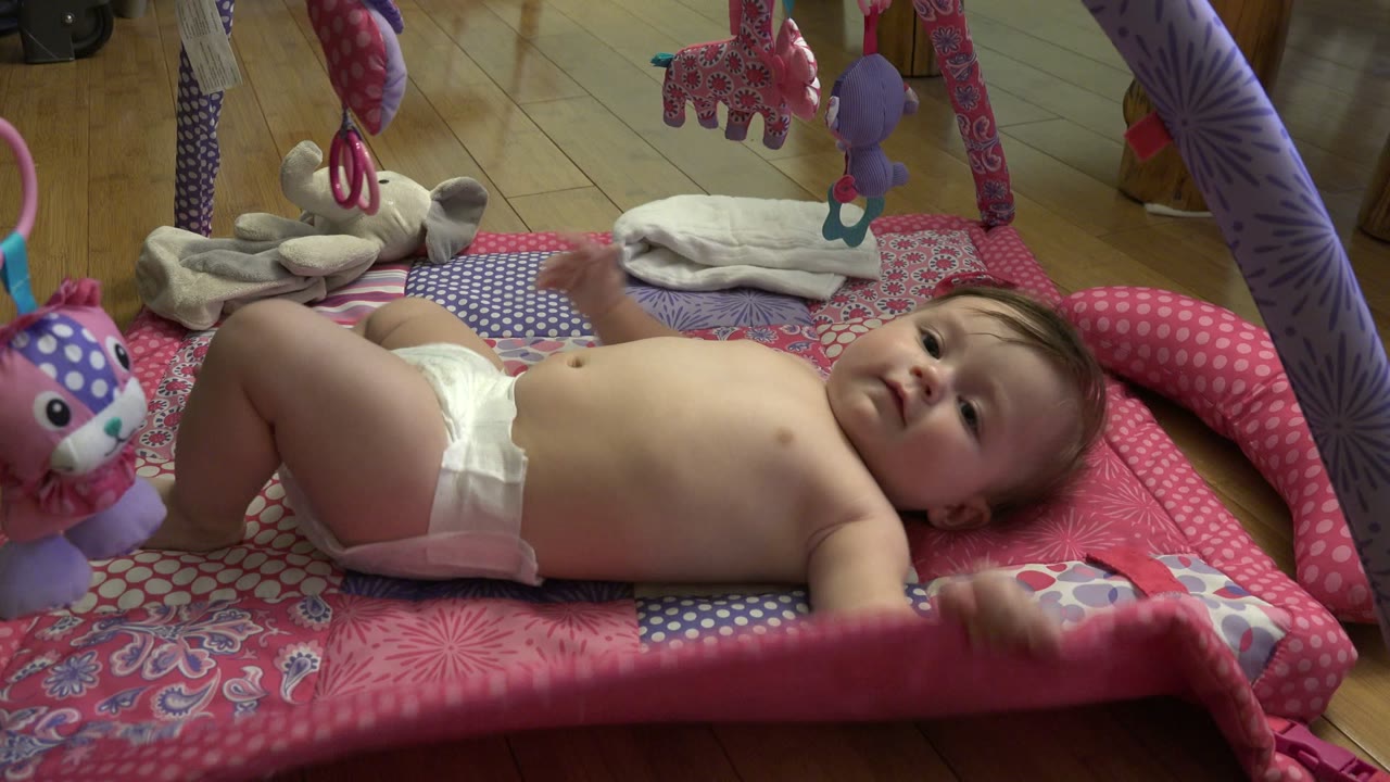 Baby lying on a pink play mat under hanging toys.