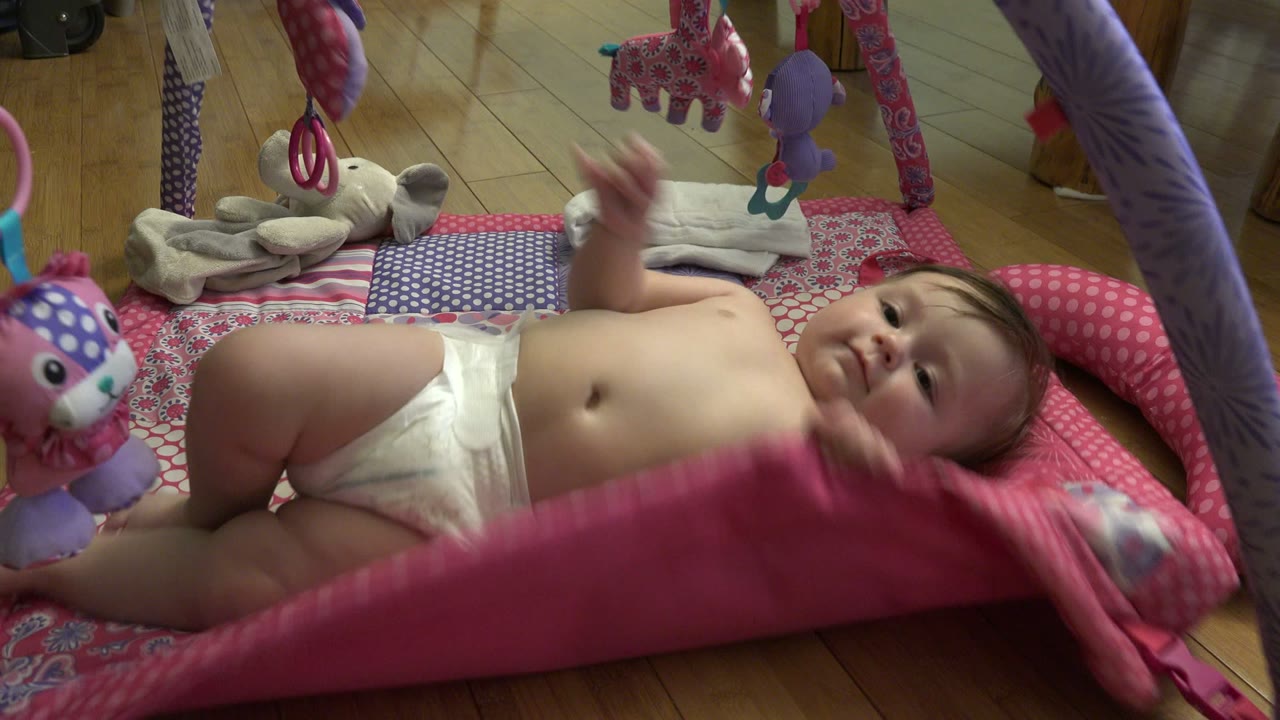 The baby remains on the patterned play mat in Westminster, Colorado, with one arm lifted toward the hanging toys and a pink edge of the mat rising in the foreground.