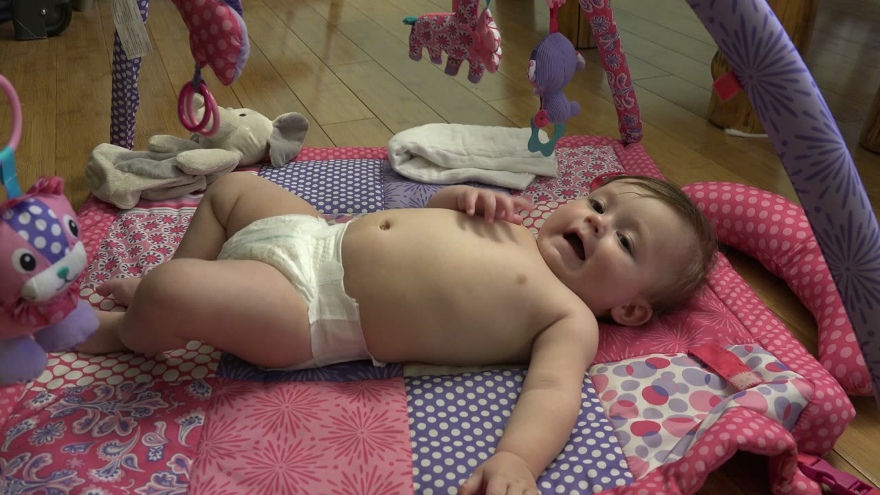 A diapered infant lies on a pink and patterned play mat under hanging baby gym toys in Westminster, Colorado.