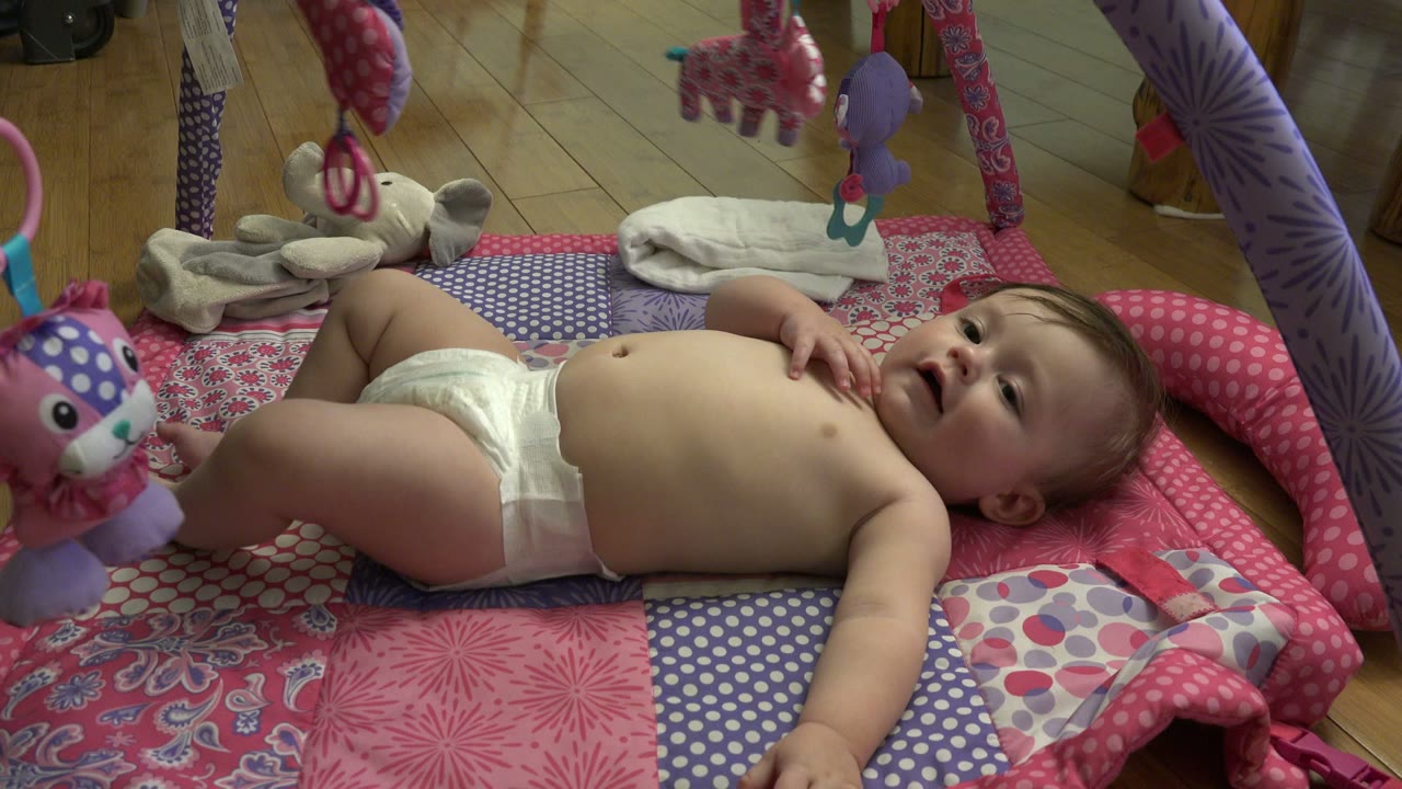 The infant remains on their back on the colorful mat, looking upward as a purple hanging toy and pink mat edge frame the Westminster home-video shot.