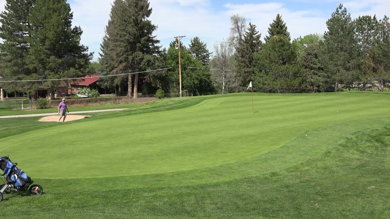 Denver golf course frame with a golf bag cart entering the lower grass area and a person in purple near the sand bunker under bright daylight.